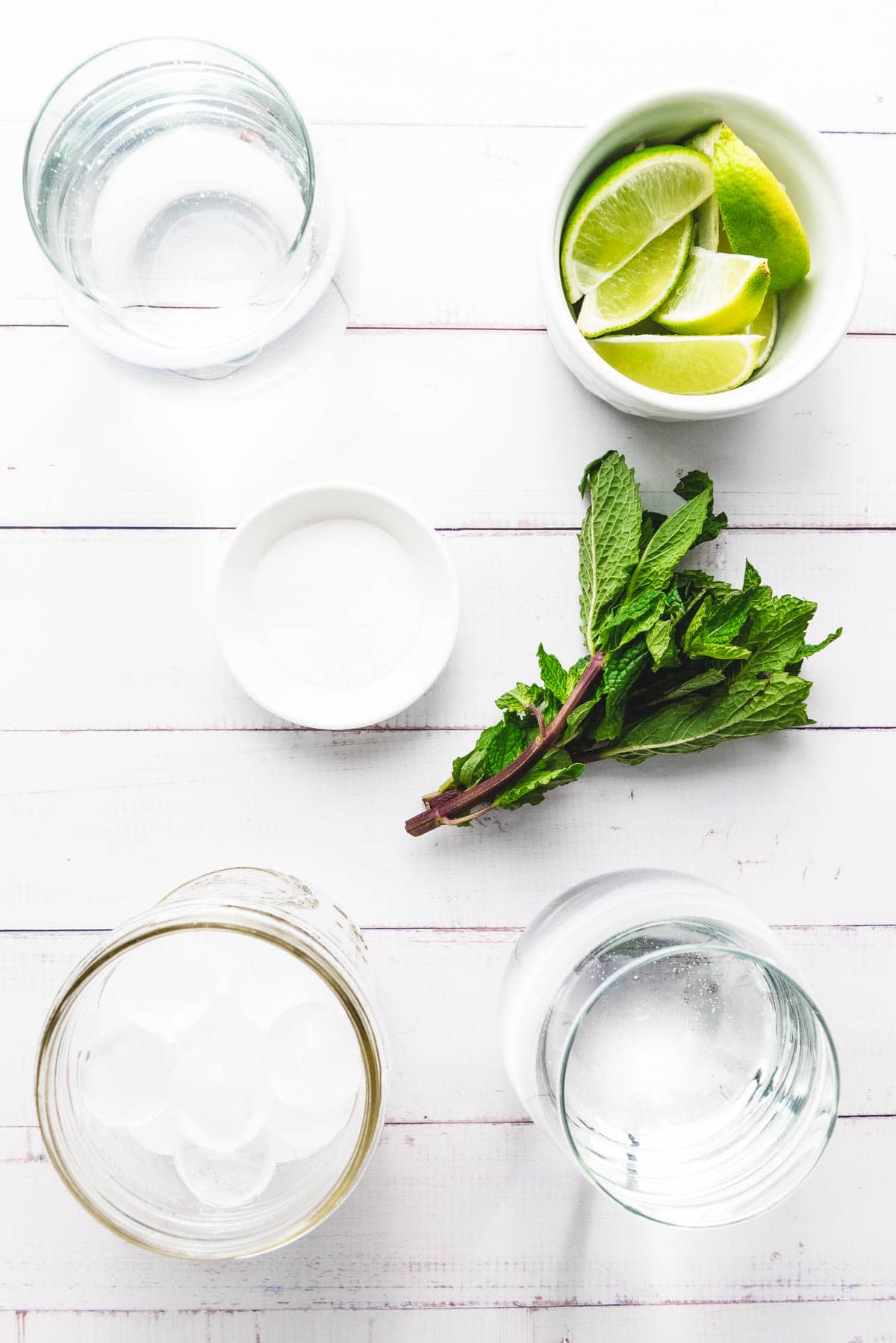 Flat lay of mint, lime wedges, ice, sugar, and water on a white wooden surface&mdash;perfect ingredients for a refreshing Faux-jito mocktail recipe.