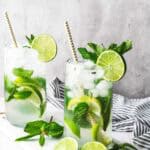 Two refreshing faux-jito cocktails with lime slices, mint leaves, and straws sit on a white surface alongside extra lime and mint—perfect for anyone seeking a delicious mocktail recipe.