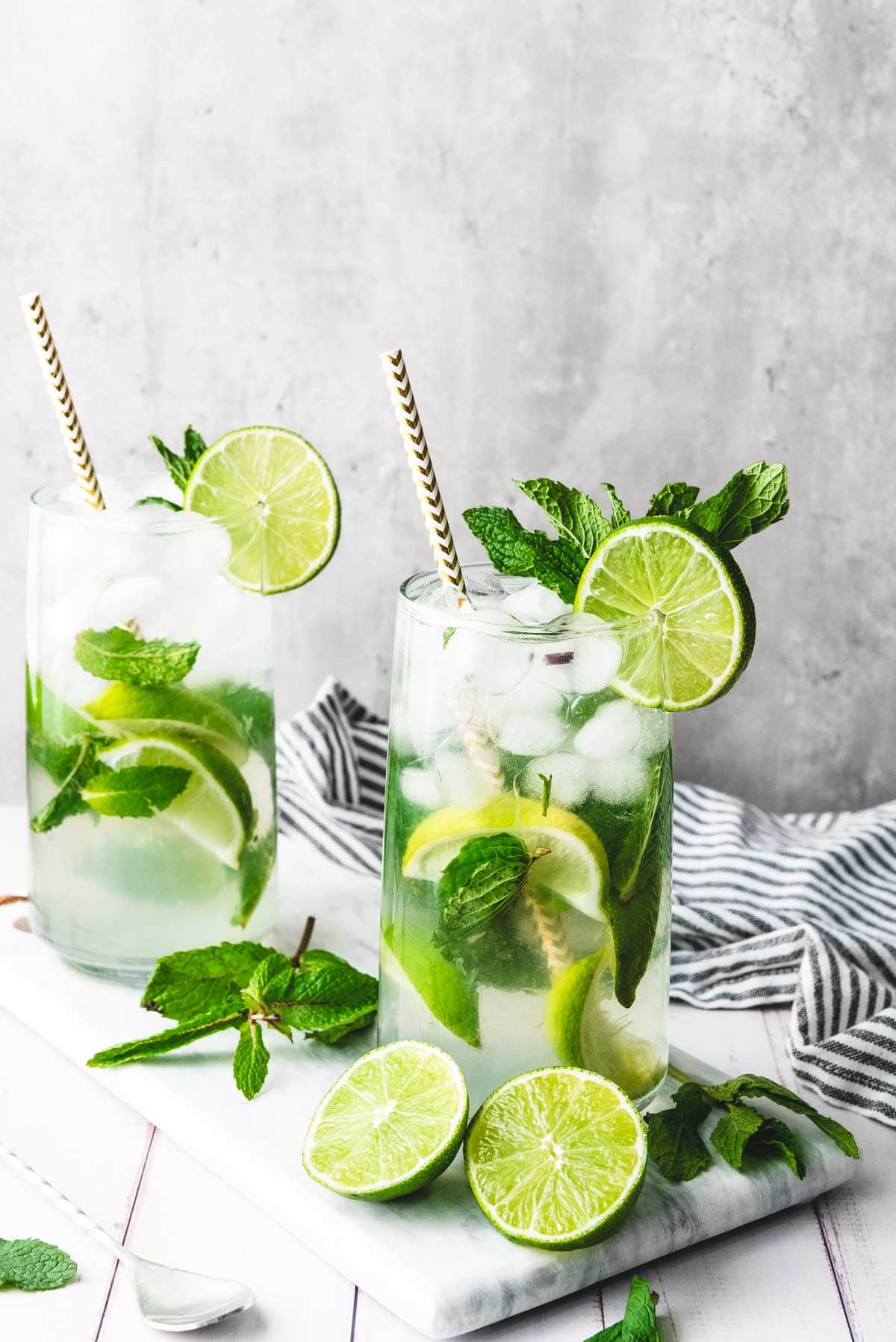 Two refreshing faux-jito cocktails with lime slices, mint leaves, and straws sit on a white surface alongside extra lime and mint&mdash;perfect for anyone seeking a delicious mocktail recipe.