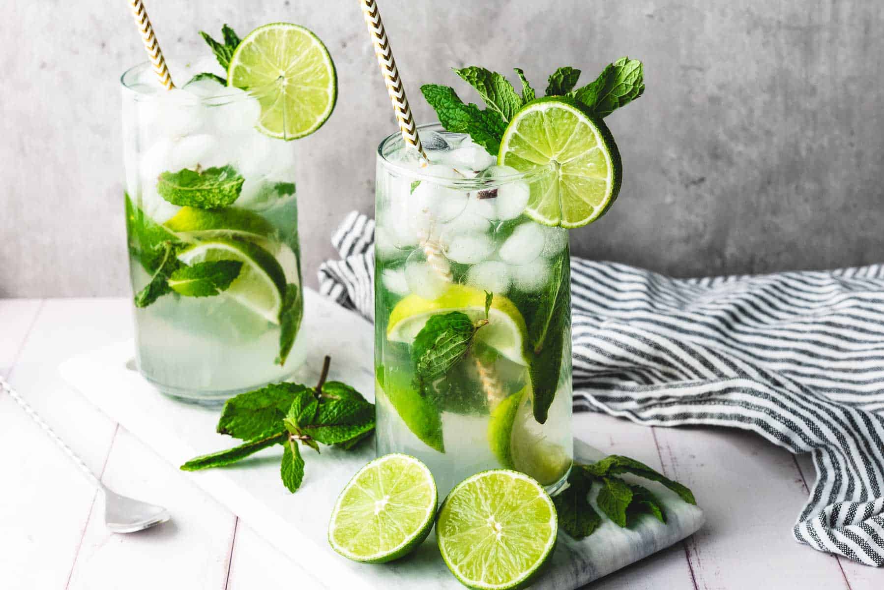 Two refreshing faux-jito cocktails with lime slices, mint leaves, ice, and straws on a white board with a striped napkin&mdash;a perfect non-alcoholic cocktail for any occasion.