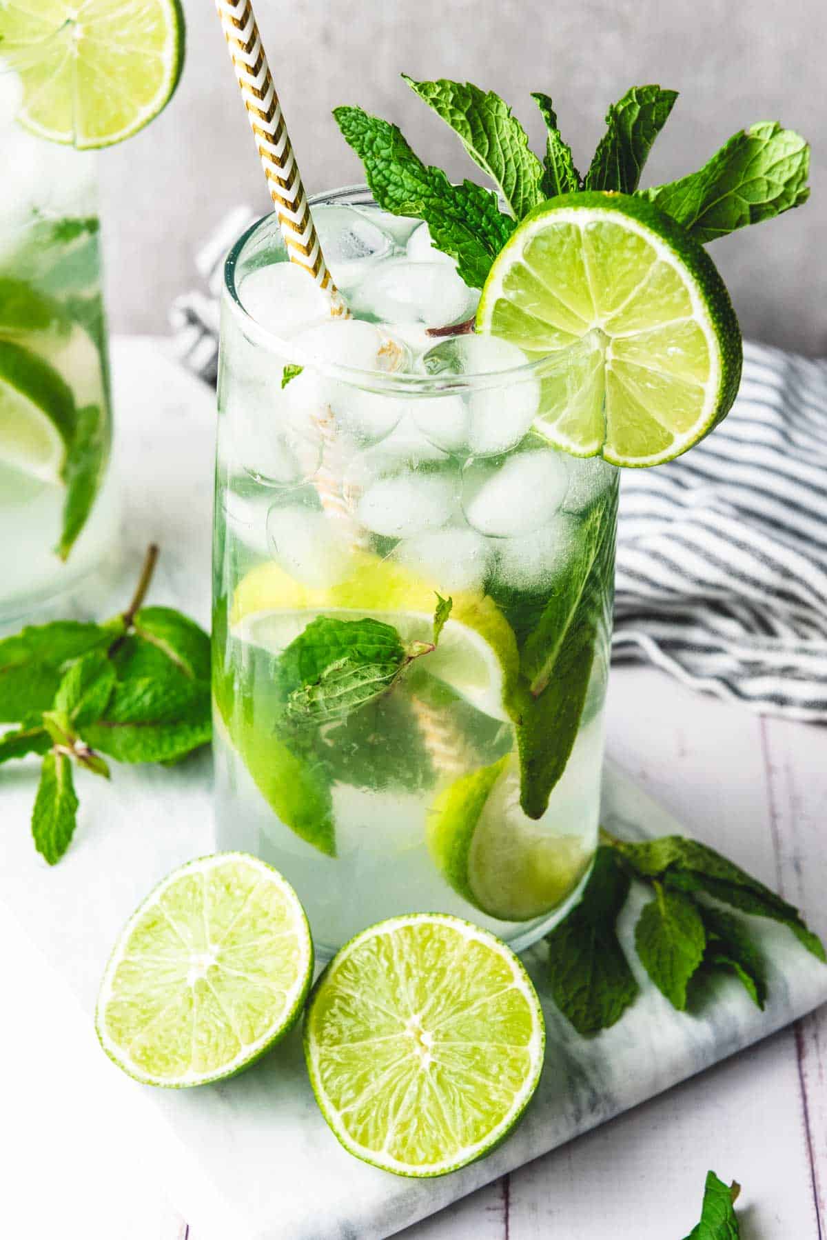 A glass of refreshing faux-jito mocktail with lime slices, mint leaves, and a striped straw, on a marble surface.