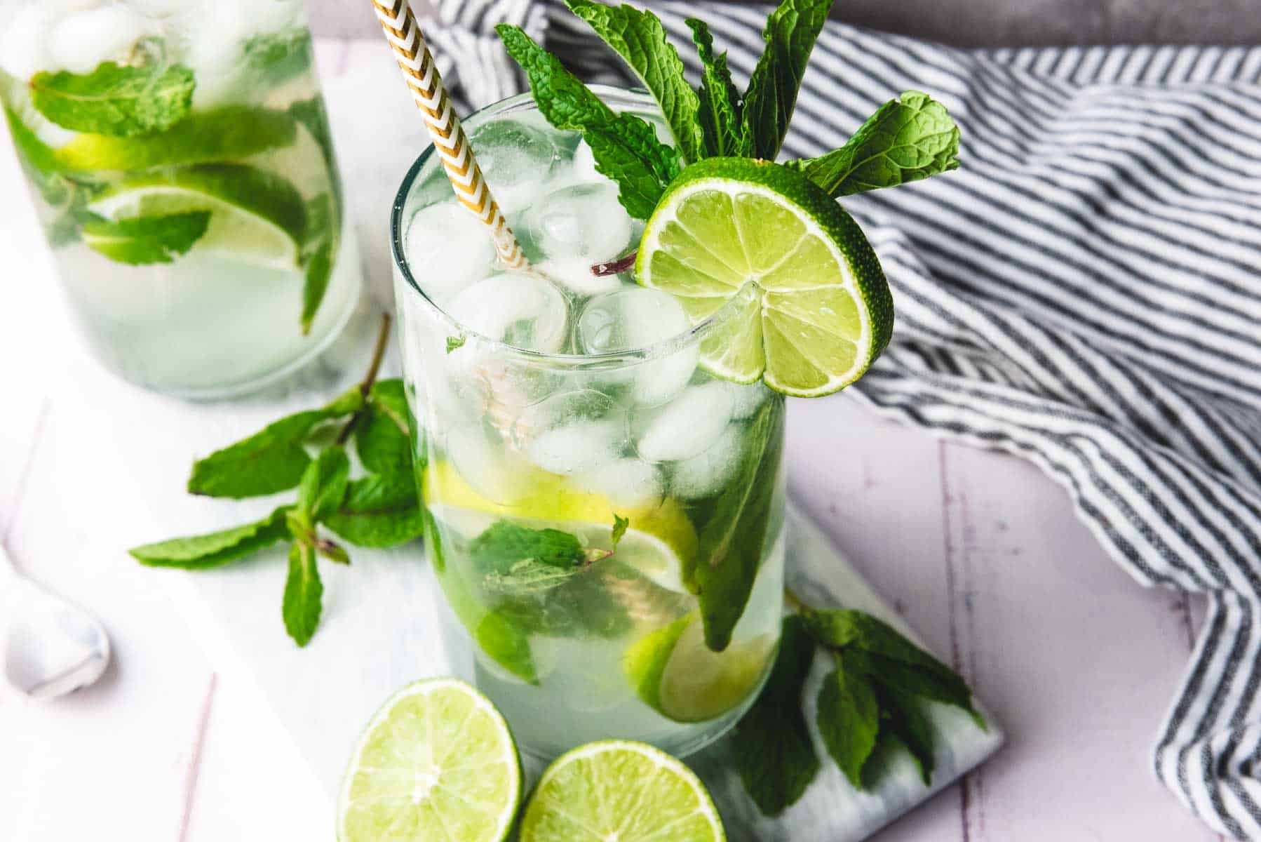 A glass of iced Faux-jito with lime slices, fresh mint, and a striped straw sits on a white surface&mdash;an inviting non-alcoholic drink for any occasion.