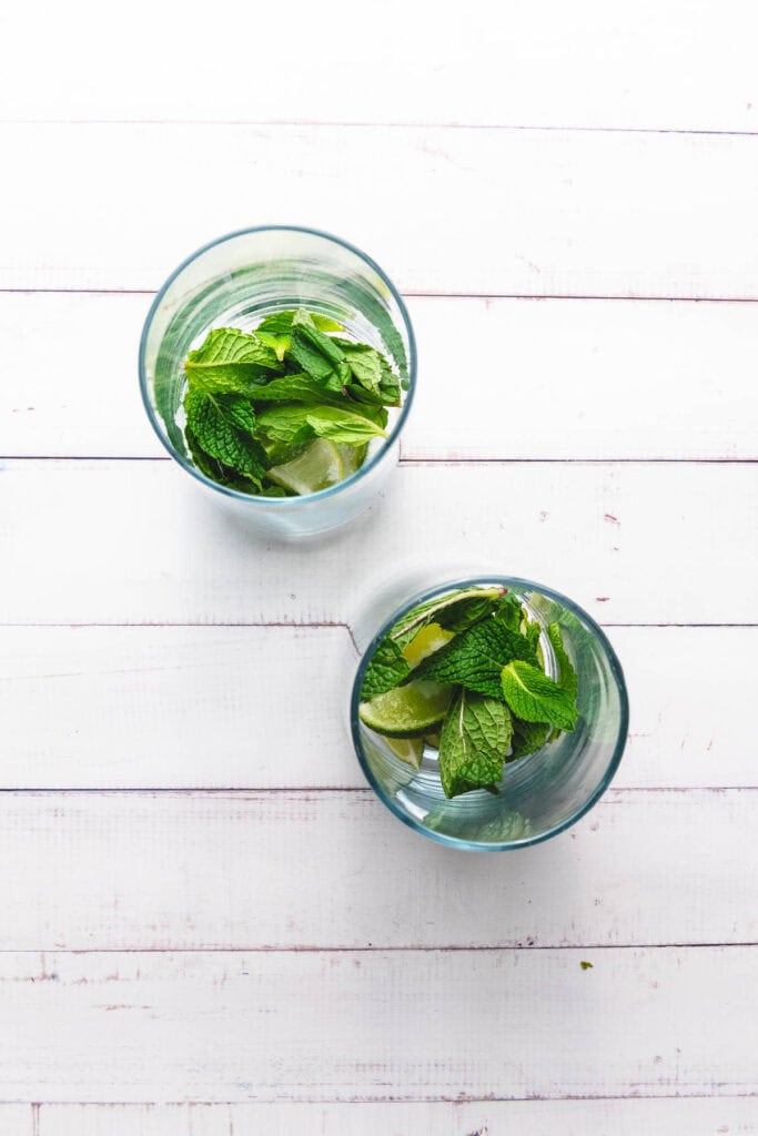 Two glasses with water, mint leaves, and lime slices on a white wooden surface, seen from above&mdash;a refreshing Faux-jito mocktail recipe that's non-alcoholic and perfect for any occasion.