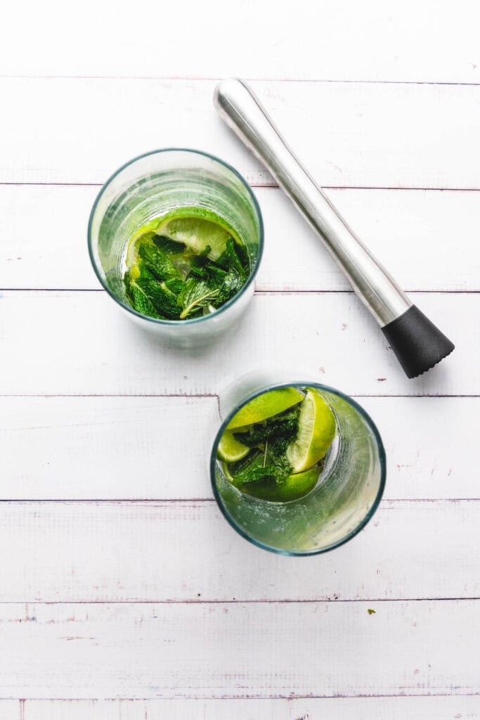 Two glasses with lime and mint, plus a muddler, on a white wooden surface&mdash;perfect for making a refreshing Mocktail or trying out your favorite Faux-jito recipe.