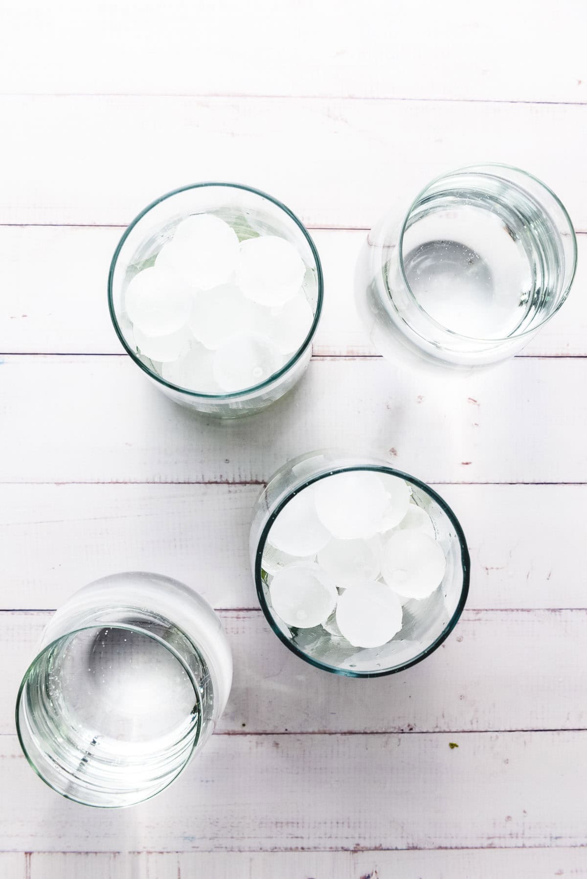 Four glasses on a white wooden surface, two filled with ice water and two with plain water&mdash;perfect for serving your favorite mocktail recipe or a refreshing Faux-jito.