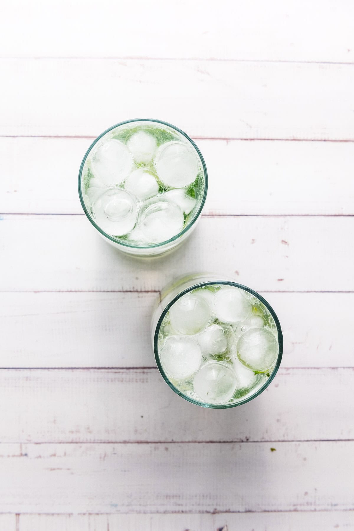 Two glasses filled with ice and a vibrant green Faux-jito mocktail sit on a white wooden surface, viewed from above.