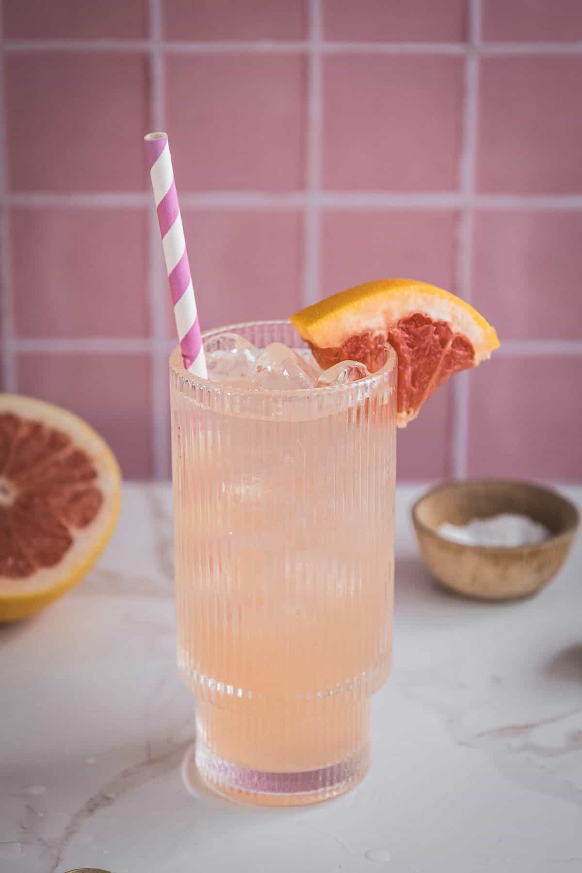 A refreshing grapefruit mocktail recipe served over ice with a striped straw and a grapefruit wedge garnish, set against a pink tiled background&mdash;perfect for an electrolyte boost.