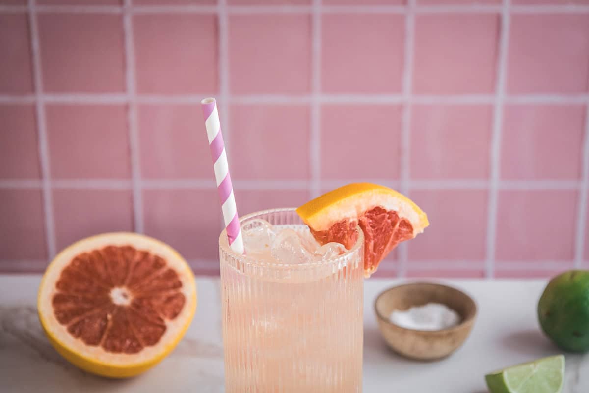 A refreshing grapefruit mocktail recipe with ice, a paper straw, and a grapefruit slice garnish, set against a pink tiled background&mdash;perfect for an electrolyte boost.