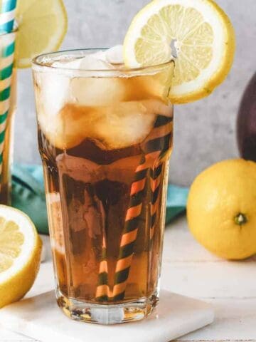 Two glasses of iced tea with lemon slices and straws, a pitcher of tea, and lemons on a white table.