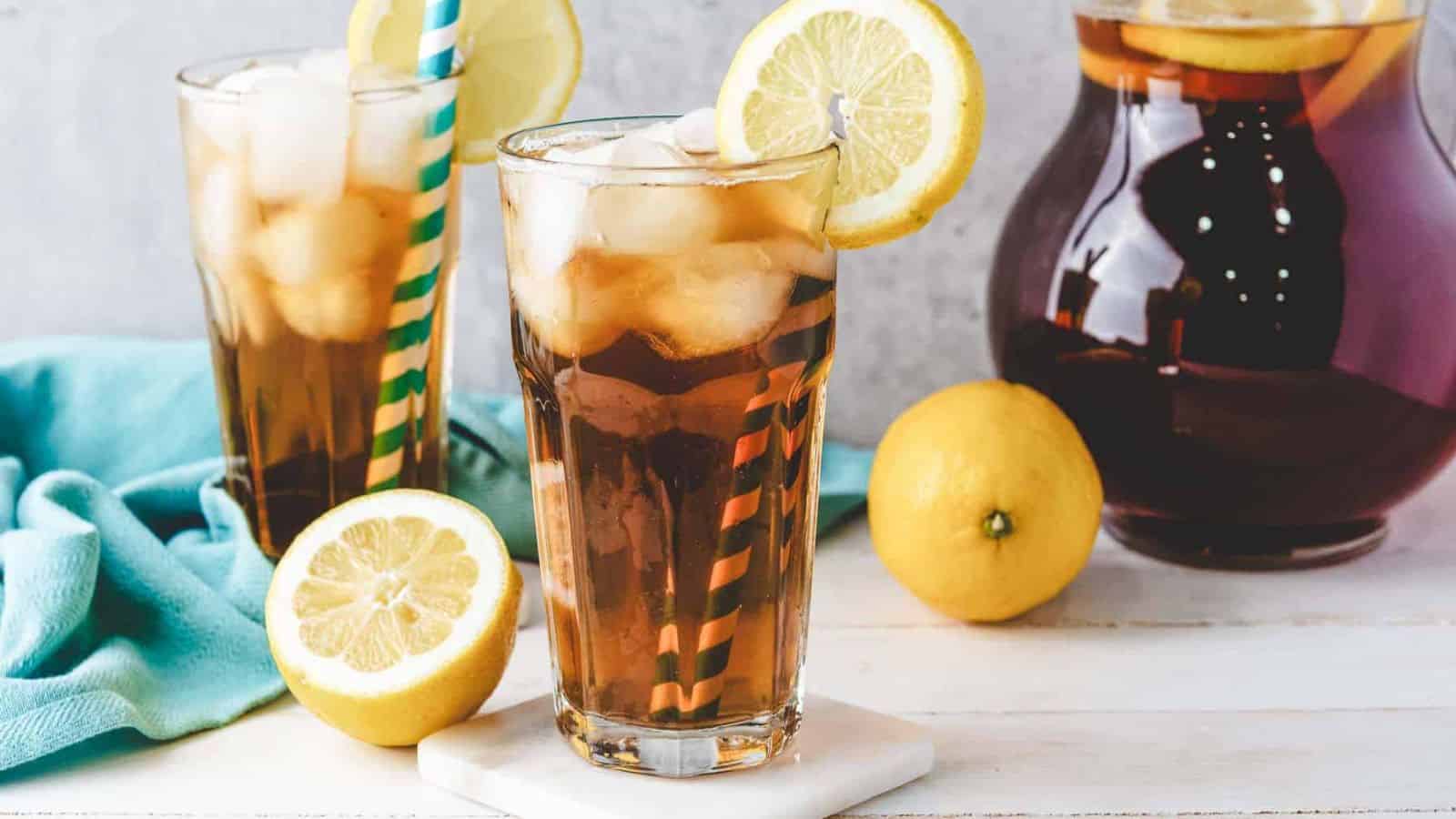 Two glasses of iced tea with lemon slices and striped straws, next to fresh lemons and a pitcher of tea.