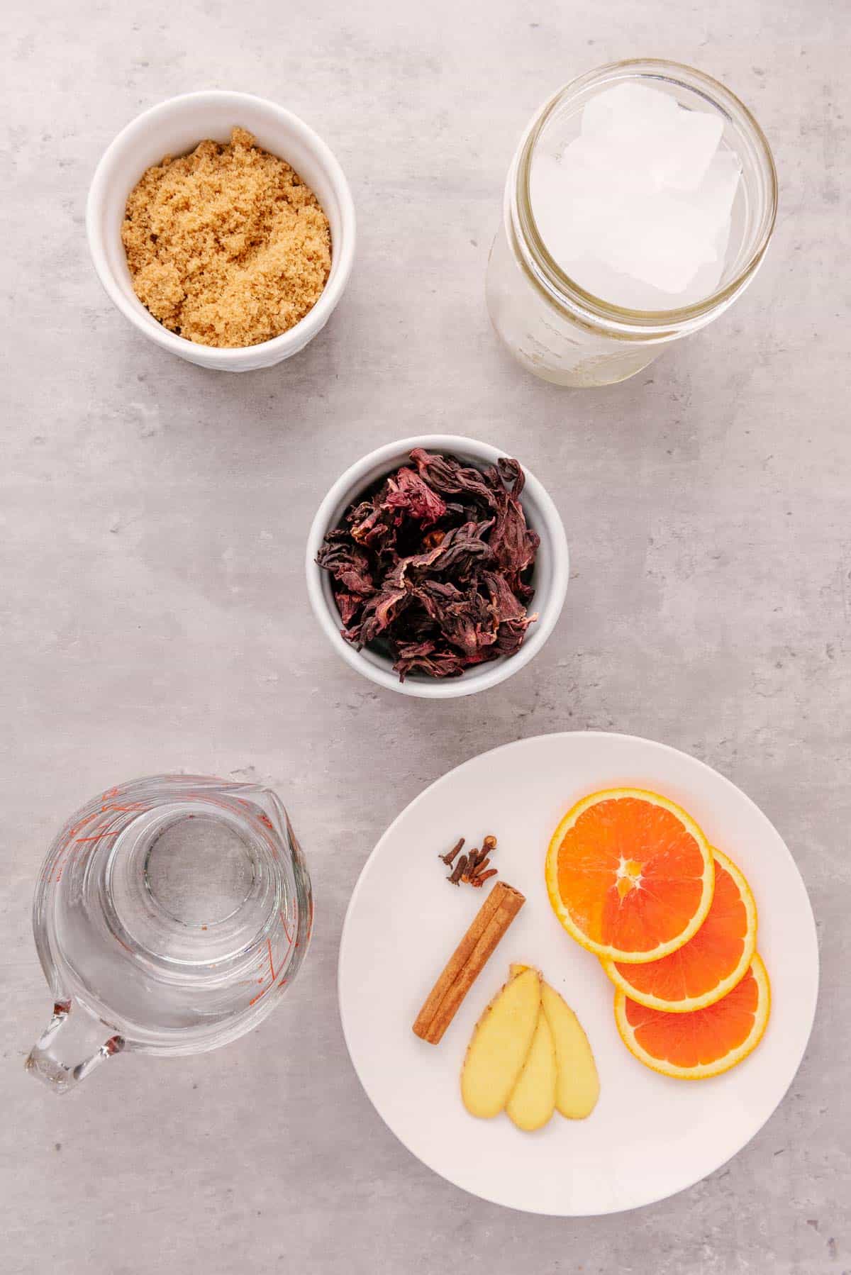 Overhead view of bowls with brown sugar, ice, dried hibiscus for making hibiscus tea, water, and a plate with orange slices, ginger, and spices&mdash;perfect ingredients for Agua de Jamaica.