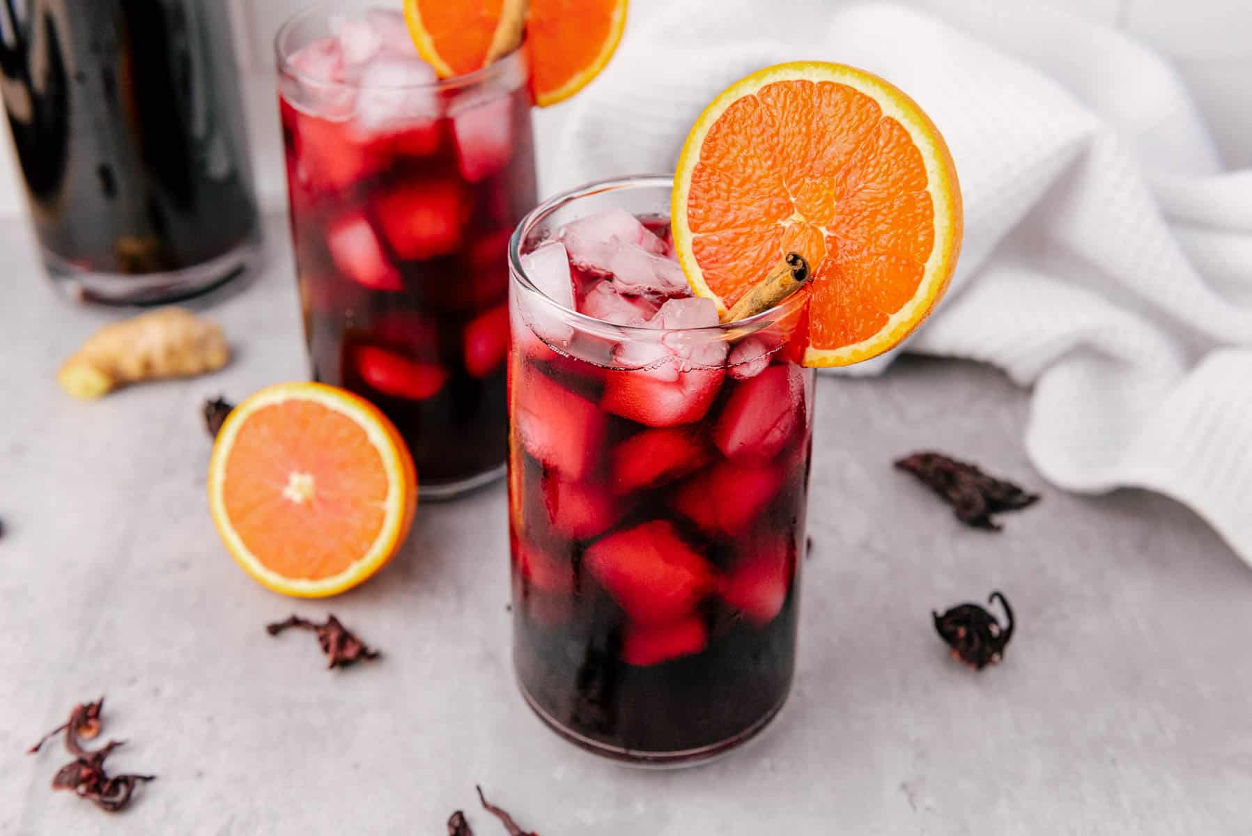 A glass of iced hibiscus tea, made from a classic Agua de Jamaica recipe, garnished with an orange slice on a light surface with scattered hibiscus petals.