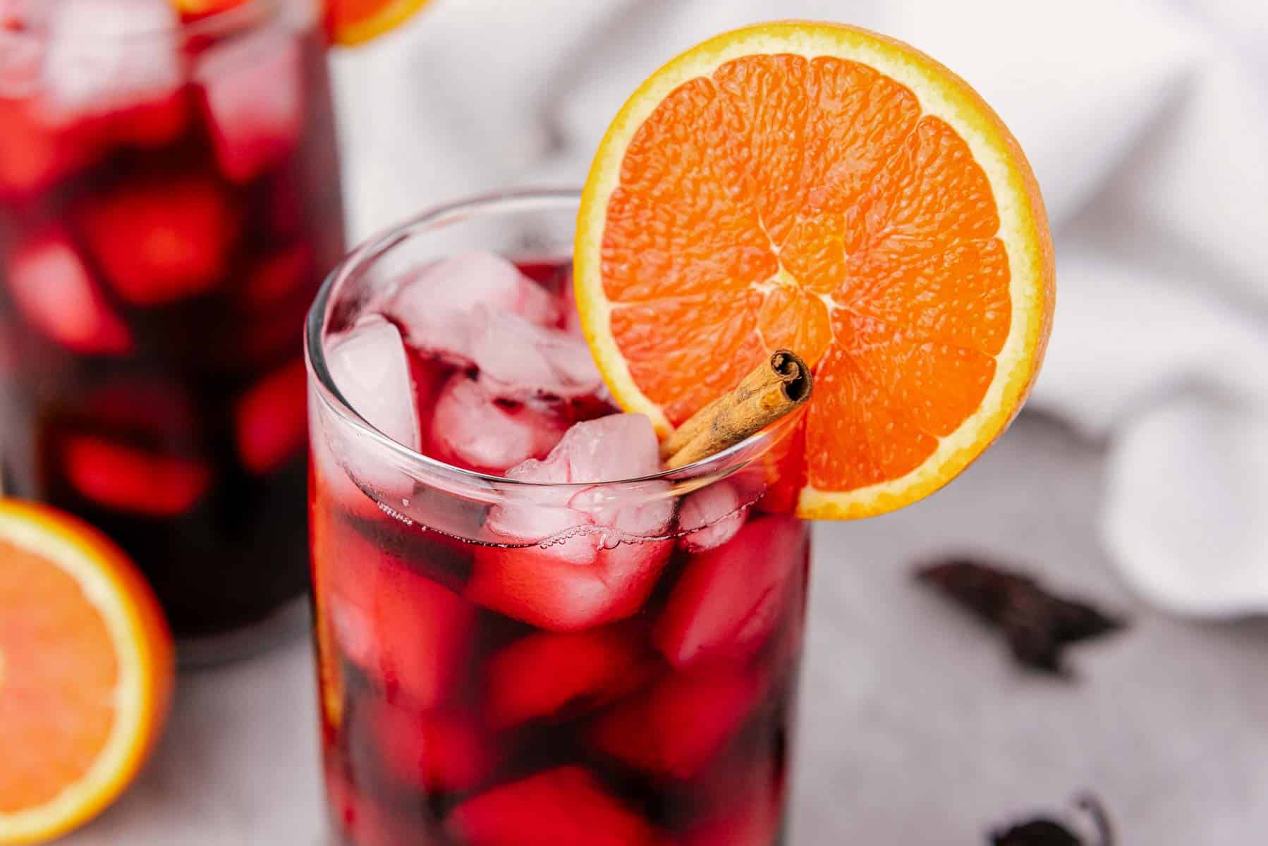 A glass of iced Jamaica drink, garnished with an orange slice and a cinnamon stick.