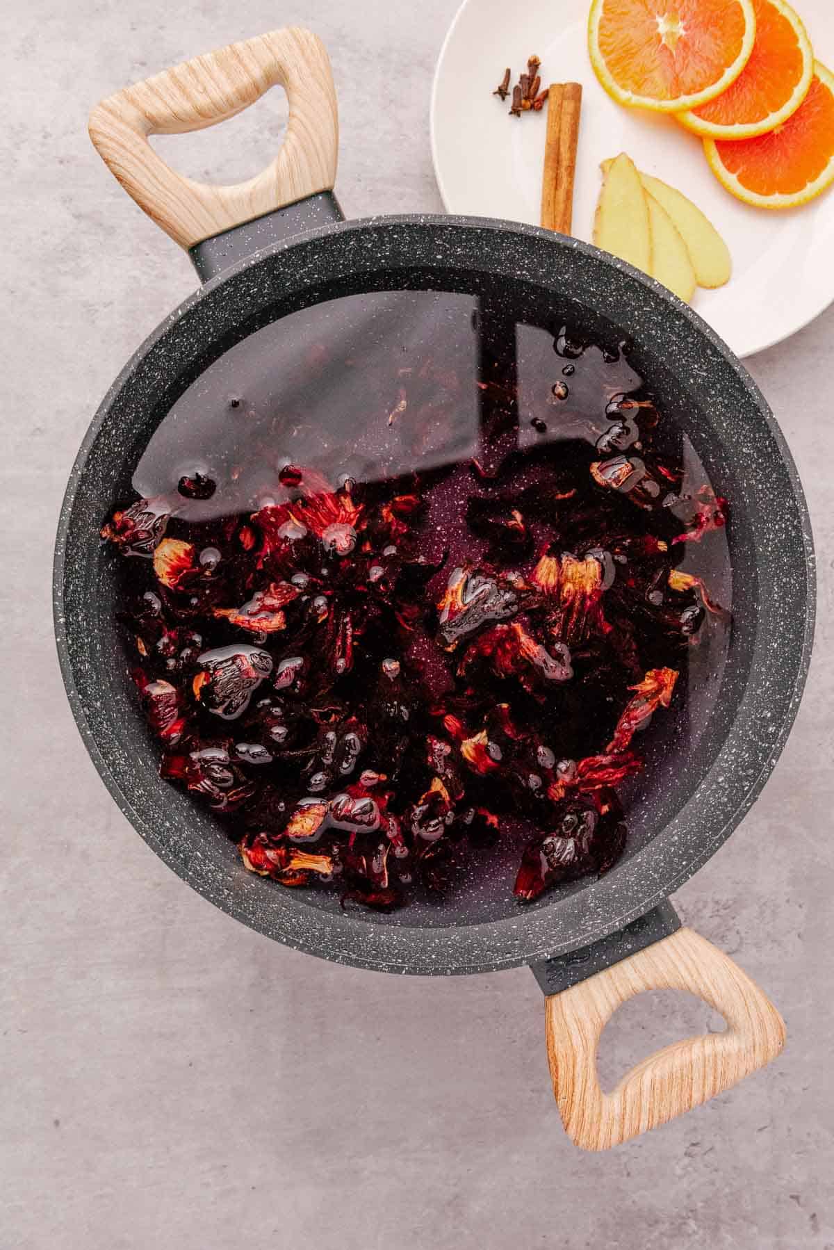 A pot of dark red hibiscus and berries in water, next to a plate with orange slices, ginger, and cinnamon&mdash;perfect ingredients for a classic hibiscus tea recipe or refreshing Agua de Jamaica.