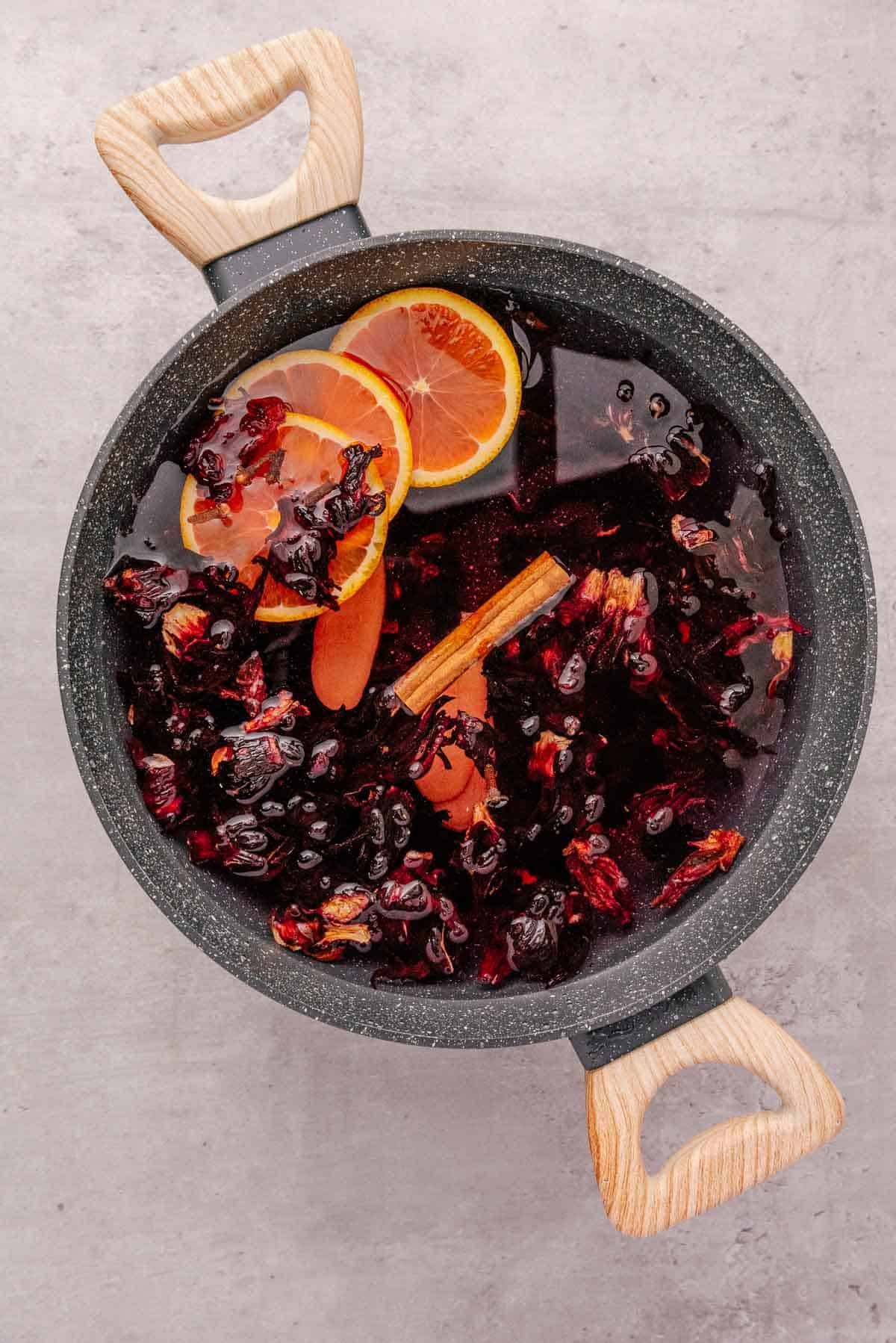 A pot of hibiscus tea, also known as Agua de Jamaica, with orange slices, cinnamon sticks, and dried hibiscus petals on a light surface.