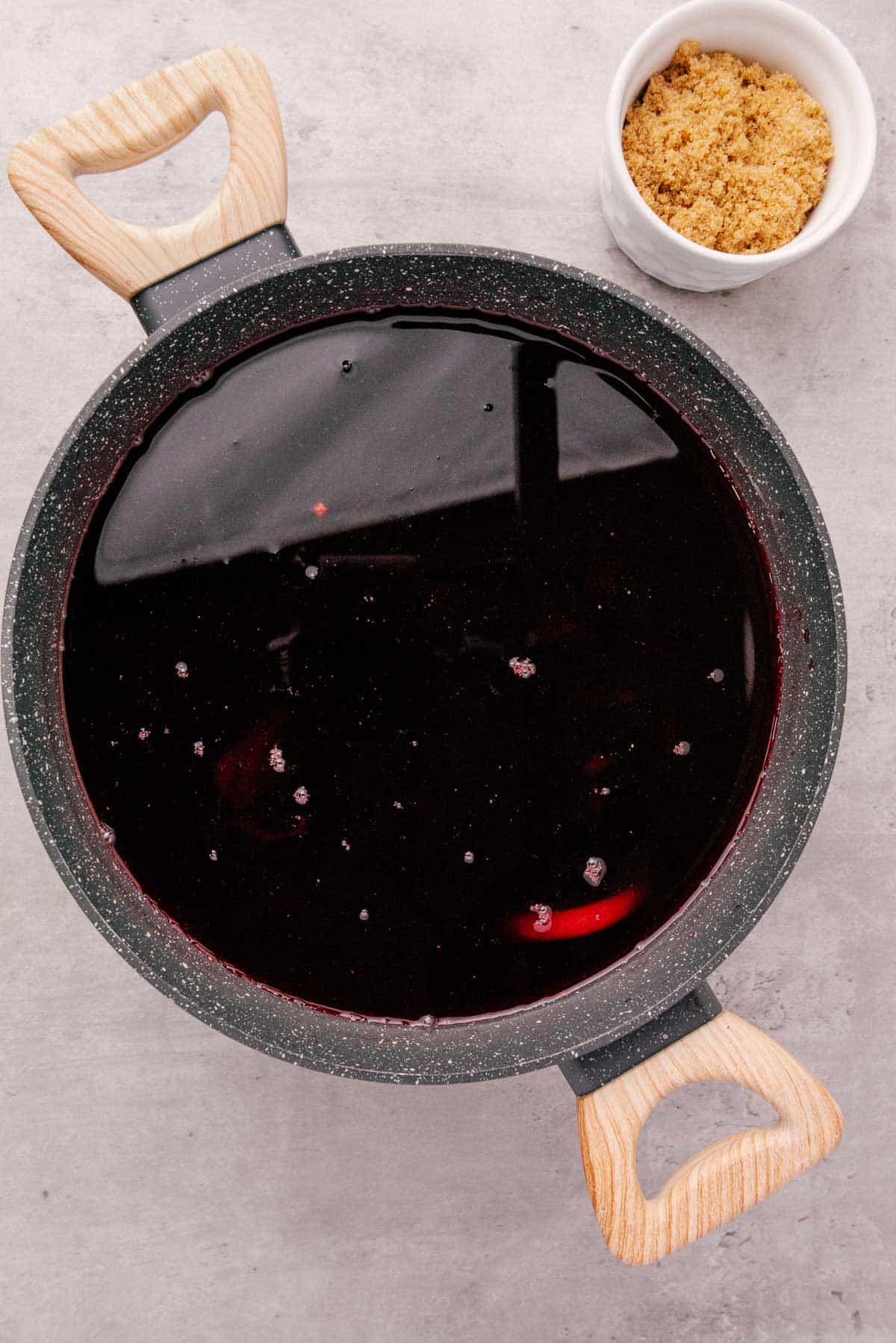 A pot of dark Jamaica drink with wooden handles sits next to a small bowl of brown sugar on a light surface.