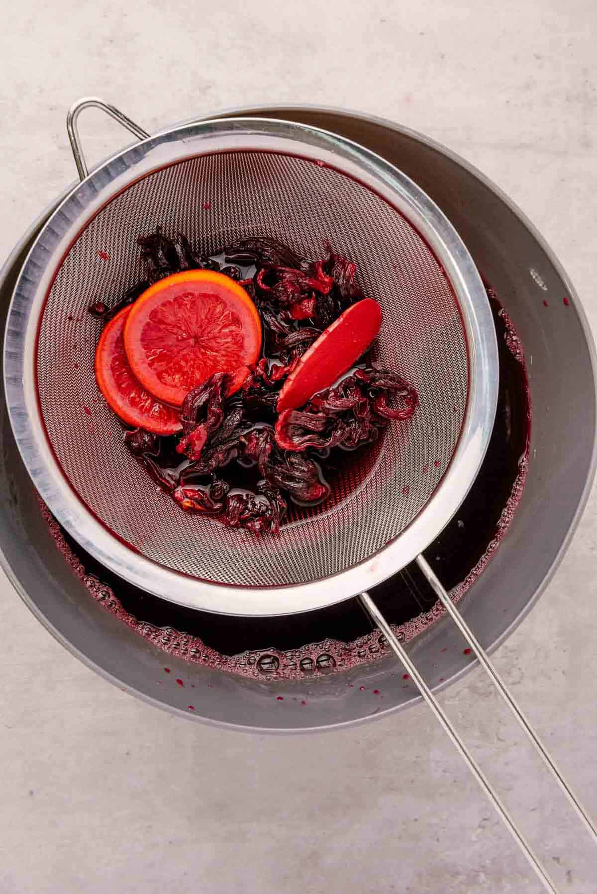 A metal strainer with hibiscus petals and orange slices rests over a bowl of dark red Agua de Jamaica, a refreshing hibiscus tea drink.