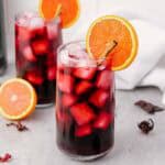 Two glasses of iced hibiscus tea, also known as Agua de Jamaica, garnished with orange slices and cinnamon sticks on a gray surface.