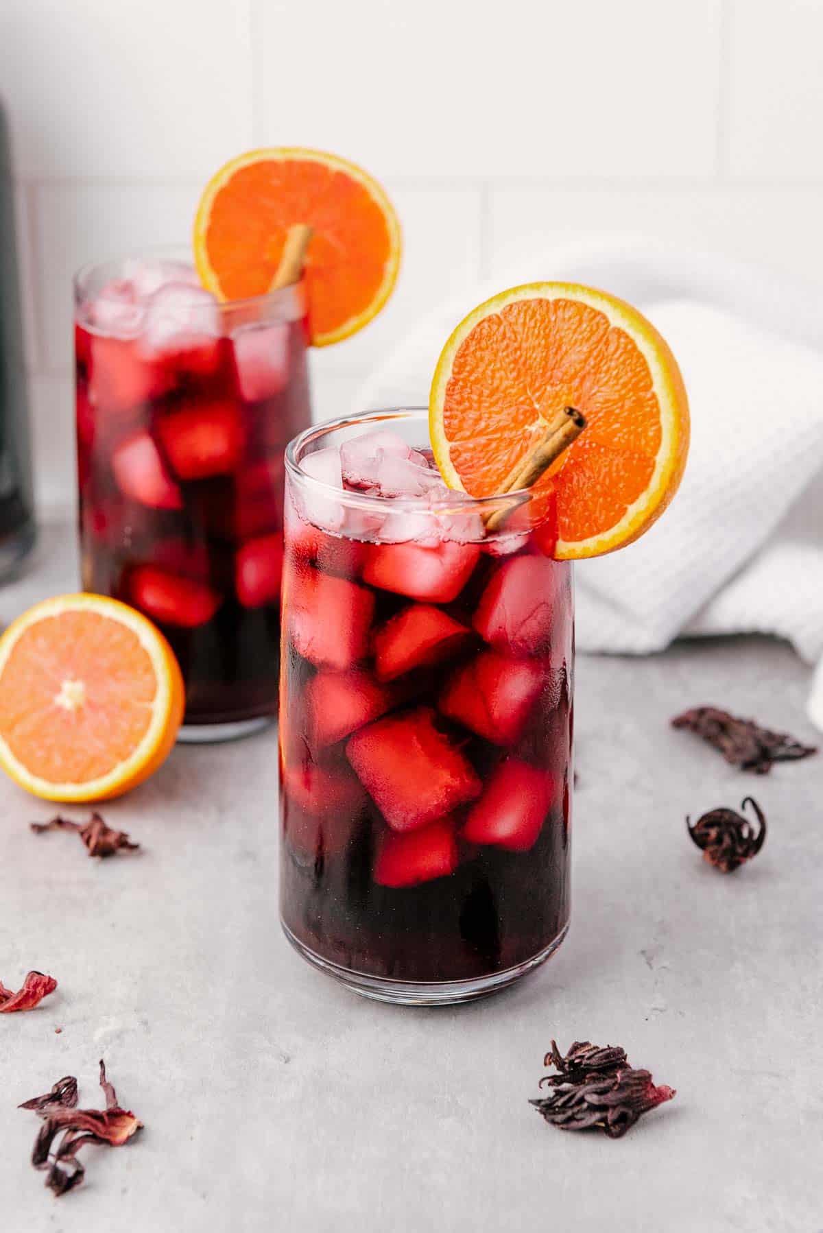 Two glasses of iced hibiscus tea, also known as Agua de Jamaica, garnished with orange slices and cinnamon sticks on a gray surface.