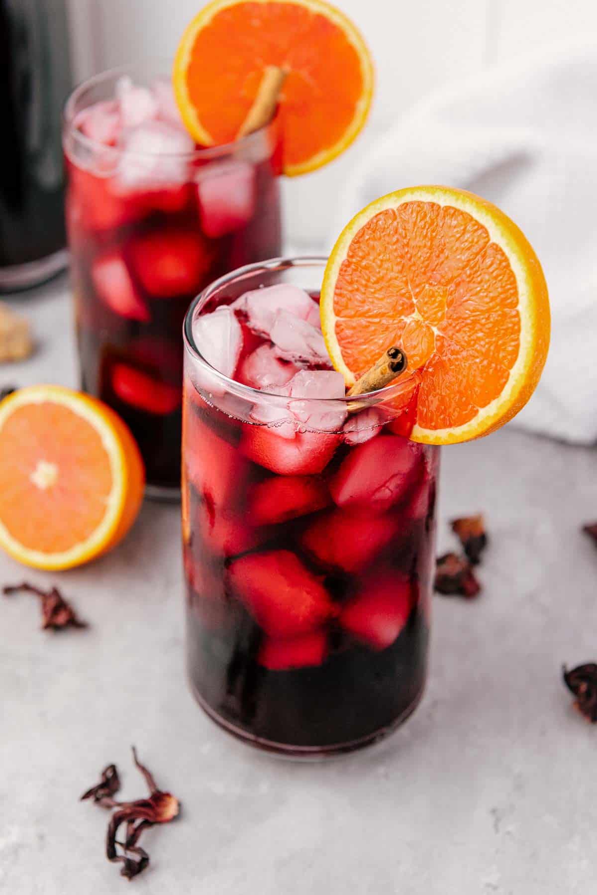 Two glasses of iced hibiscus tea, also known as Agua de Jamaica, garnished with orange slices on a gray surface.