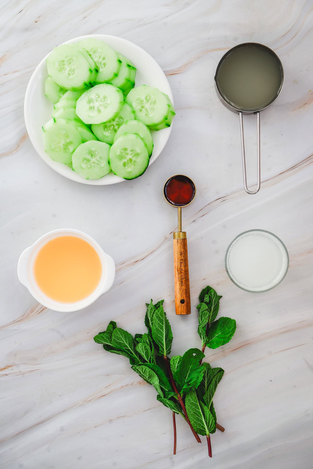 Sliced cucumbers and mint leaves sit beside liquids in bowls and a cup on a white marble surface&mdash;perfect for crafting a refreshing Margarita Mocktail recipe.