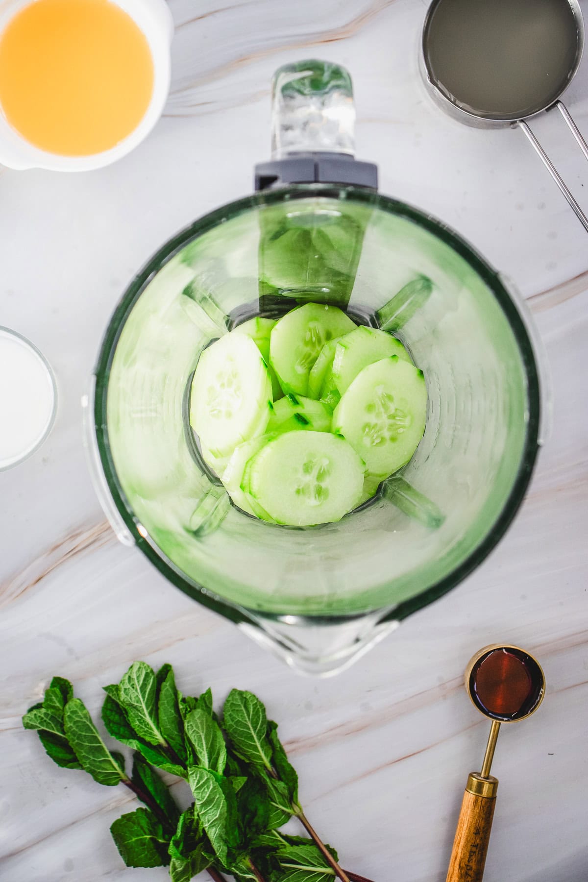 Sliced cucumber in a blender surrounded by mint, a measuring spoon, and bowls of liquids on a white surface&mdash;perfect for making a refreshing Cucumber Margarita Mocktail.