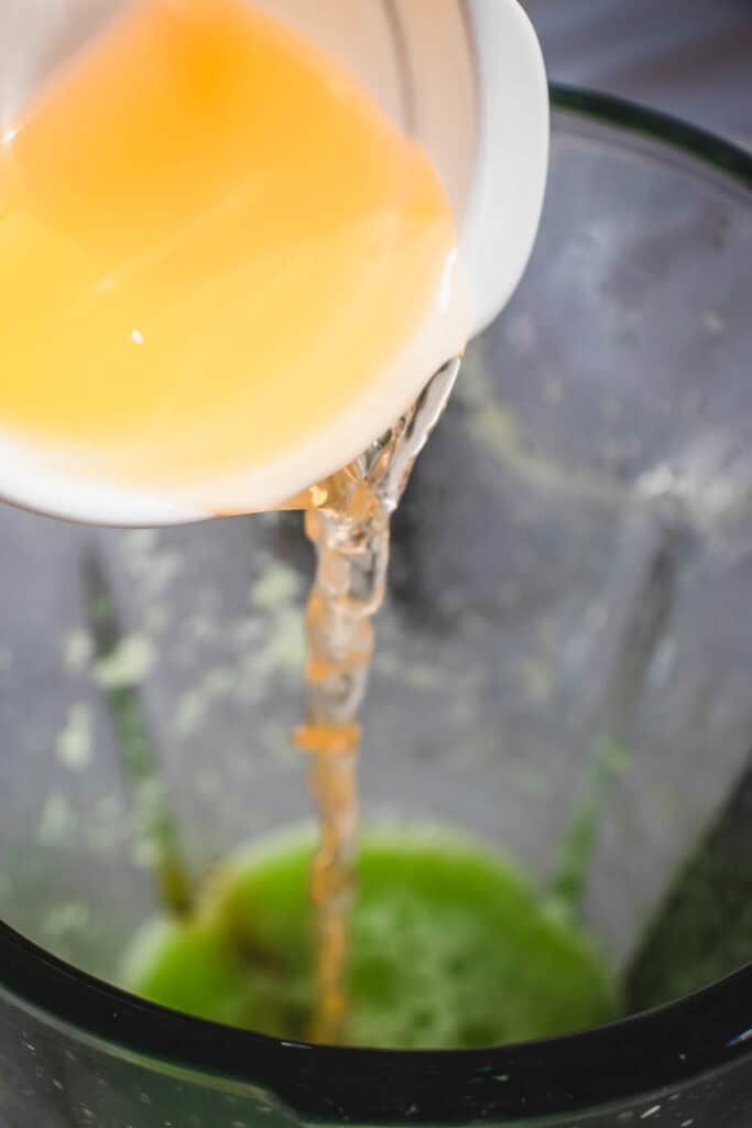 A bowl of orange liquid is poured into a blender containing a green cucumber mixture, creating a vibrant Margarita Mocktail.