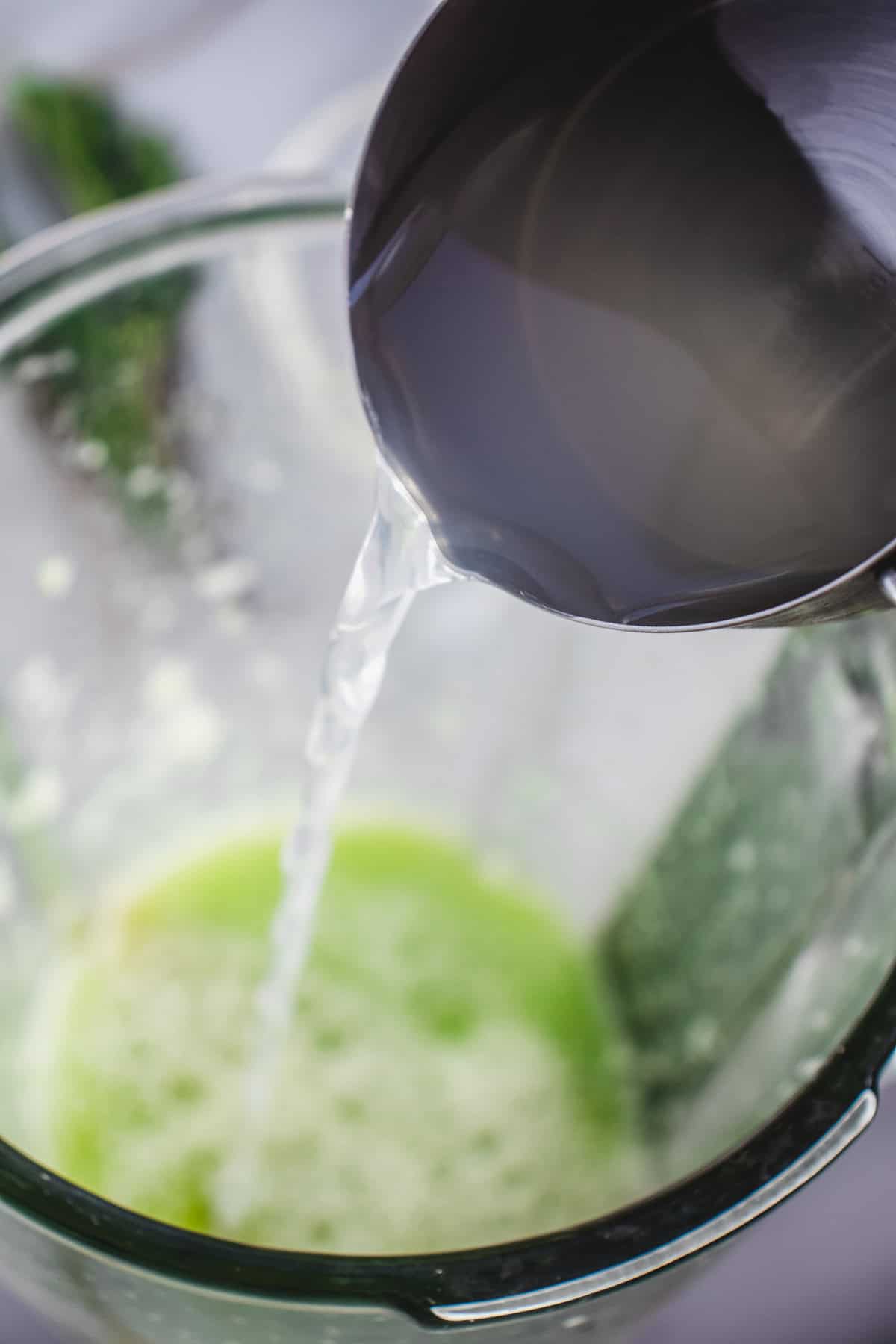 Water being poured from a metal container into a blender with a green mixture inside, perfect for crafting a refreshing Cucumber Mocktail.