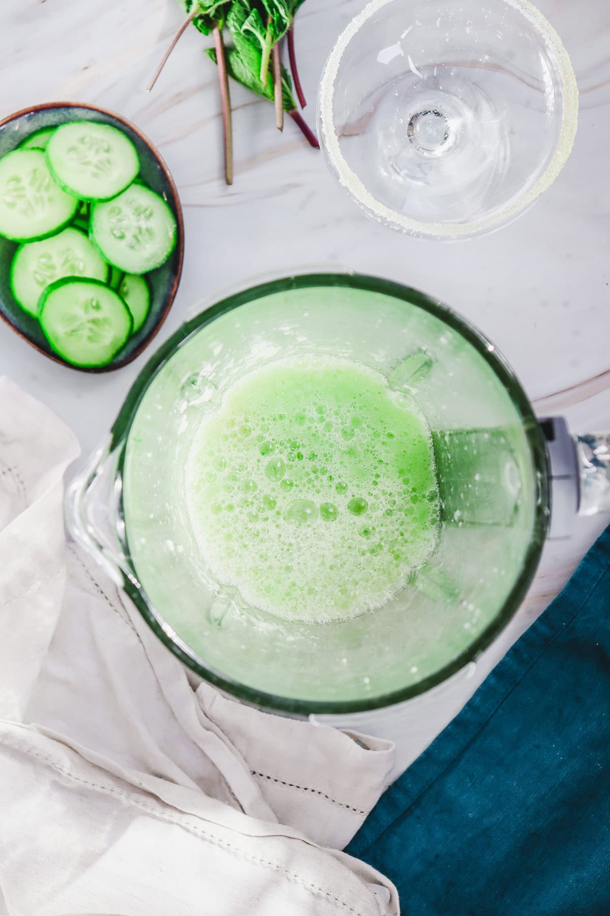 Top view of a blender with a green smoothie, cucumber slices, and a glass on a white and blue surface&mdash;perfect for creating a refreshing Cucumber Margarita Mocktail.