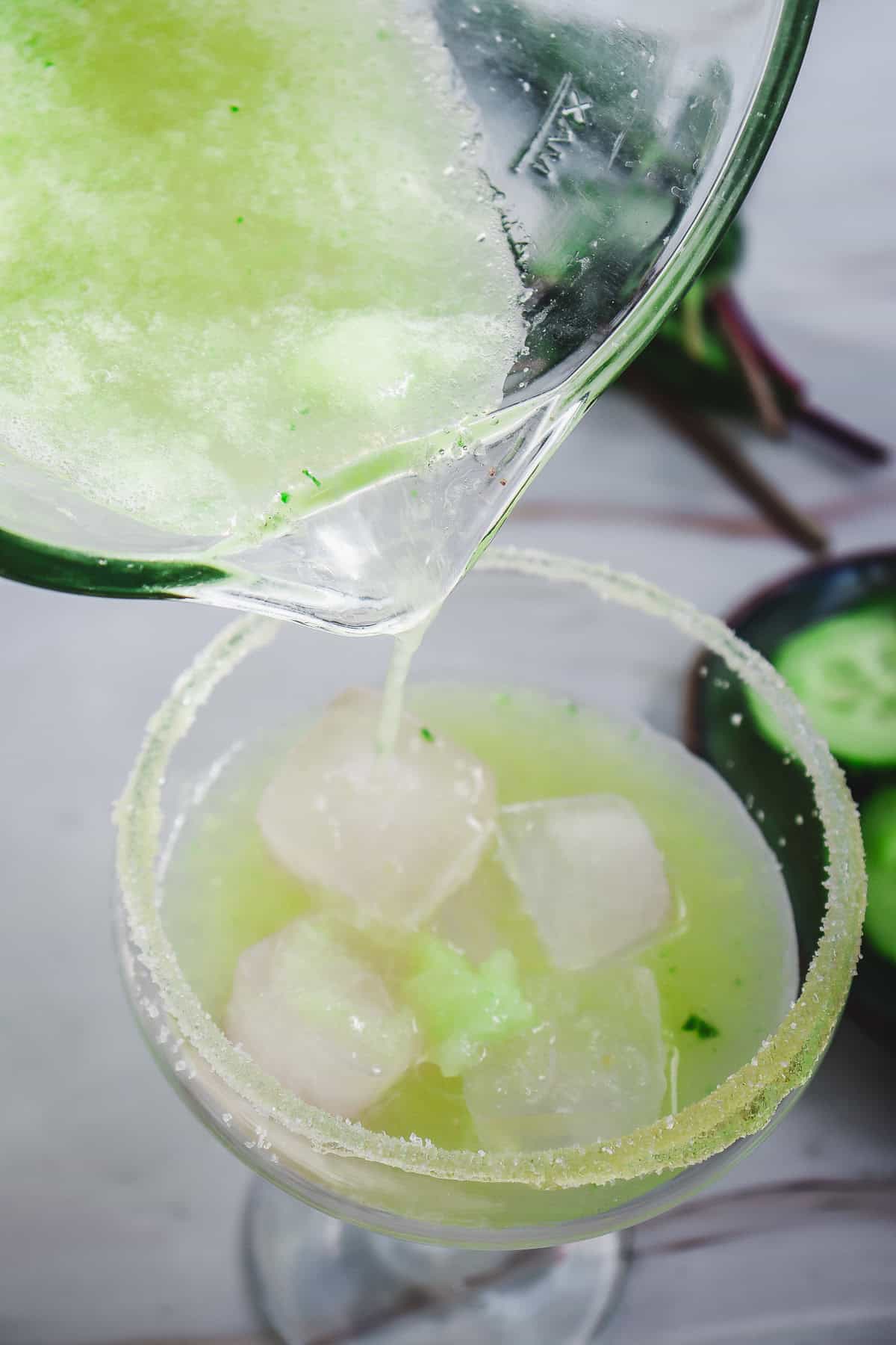 A vibrant green Cucumber Margarita Mocktail is being poured into a sugar-rimmed glass filled with ice cubes.