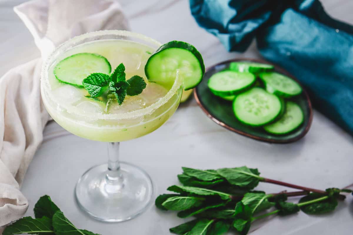 A refreshing Cucumber Margarita Mocktail with ice, cucumber slices, and mint garnish in a glass, with fresh mint and cucumber nearby.
