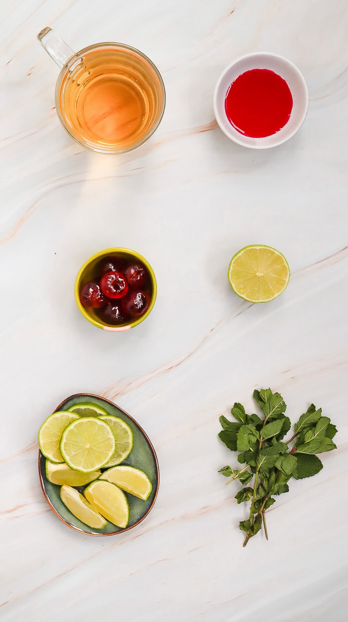Glass of juice, bowl of red liquid, cherries, lime halves, lime slices, and mint on a marble surface&mdash;perfect ingredients for a refreshing Shirley Temple mocktail recipe.