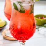 A red fizzy Shirley Temple mocktail with ice, mint, lime slice, and cherries in a curved glass on a woven coaster.