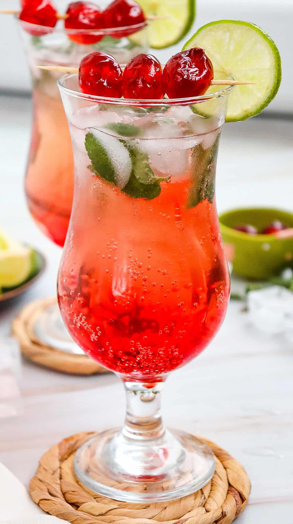 A red fizzy Shirley Temple mocktail with ice, mint, lime slice, and cherries in a curved glass on a woven coaster.