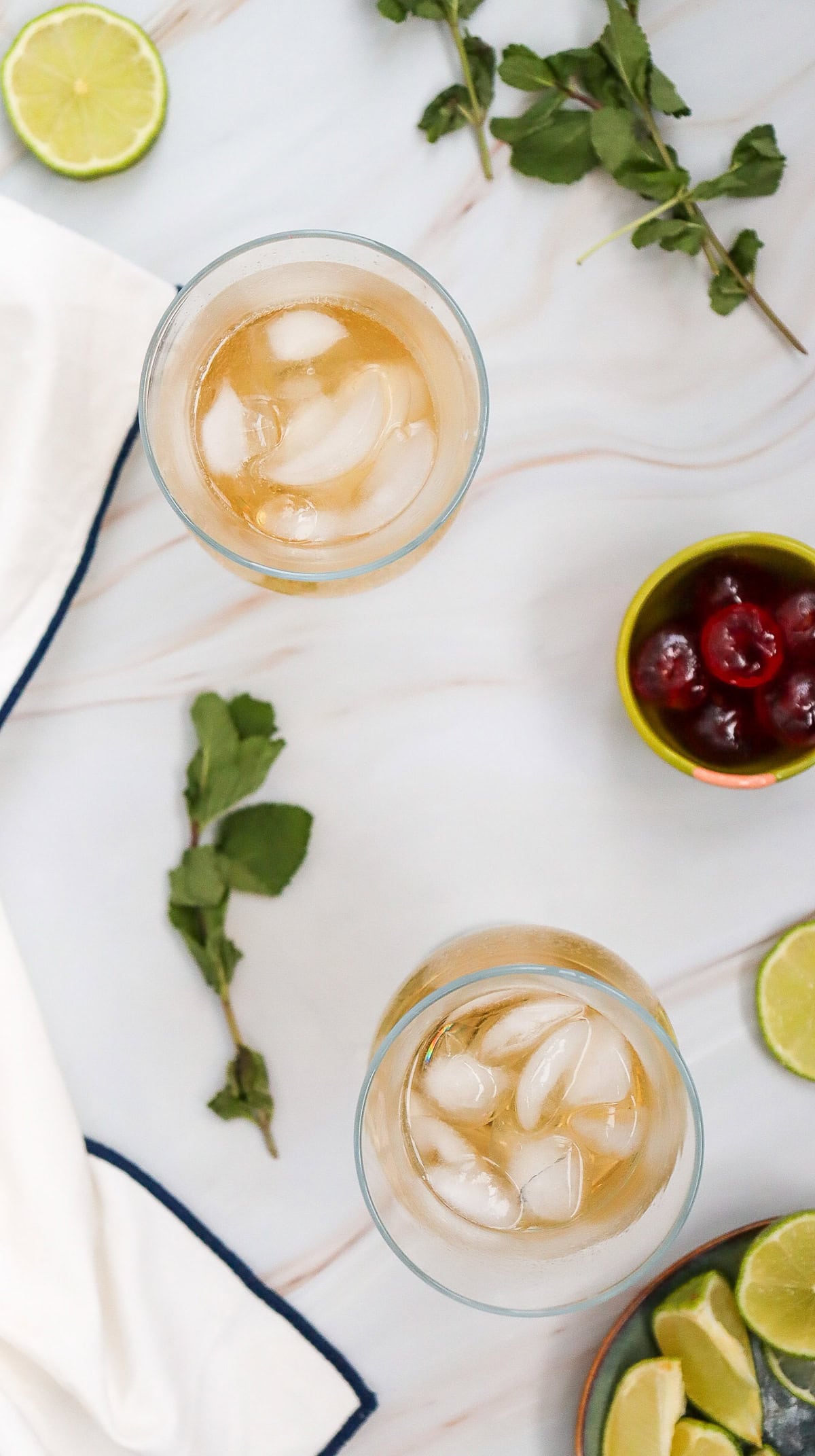 Two glasses of iced Shirley Temple mocktail on a white surface, garnished with lime slices, mint leaves, and cherries arranged around them&mdash;perfect for trying a refreshing new recipe.