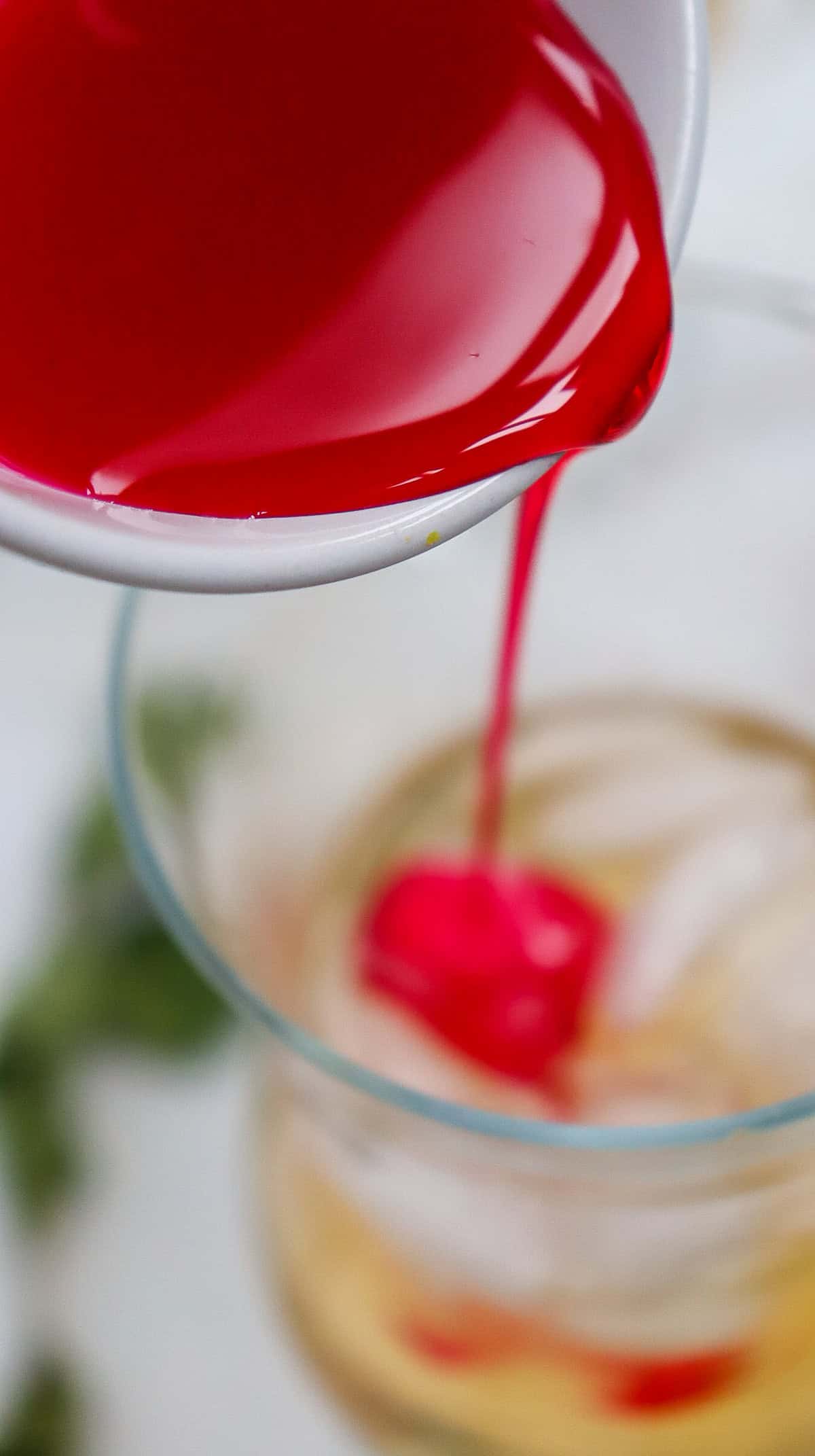 Red liquid being poured from a white bowl into a glass with ice, creating the perfect Shirley Temple Mocktail, with a blurred background.