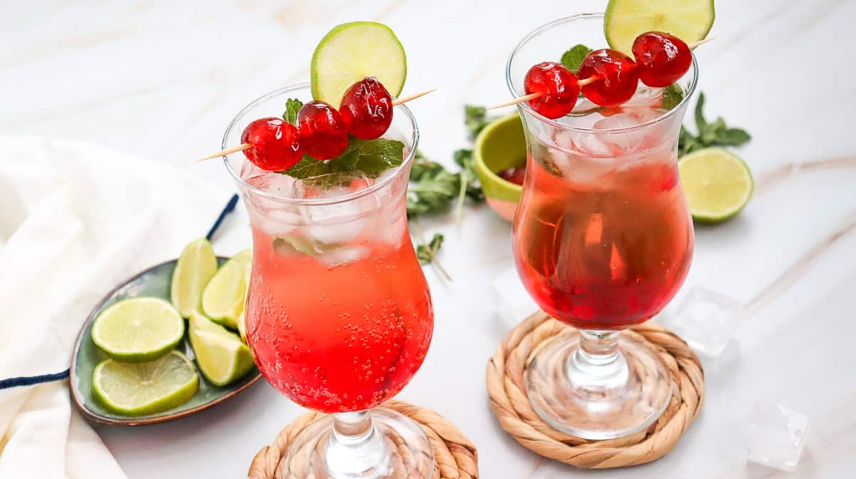 Two iced red Shirley Temple Mocktails in tall glasses, garnished with lime, mint, and cherries sit on a white table alongside sliced limes&mdash;a refreshing mocktail recipe perfect for any occasion.