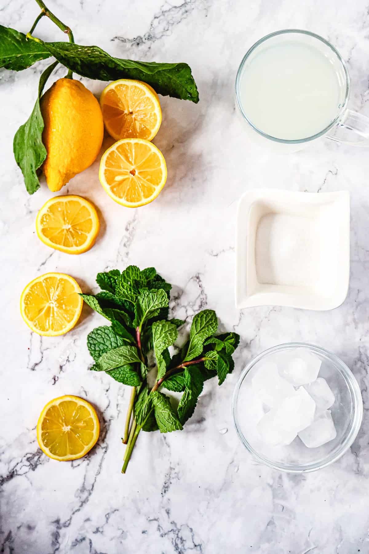 Lemon, lemon slices, mint, ice cubes, sugar, and lemon juice arranged on a marble surface&mdash;perfect ingredients for a refreshing Lemon Mojito Recipe.