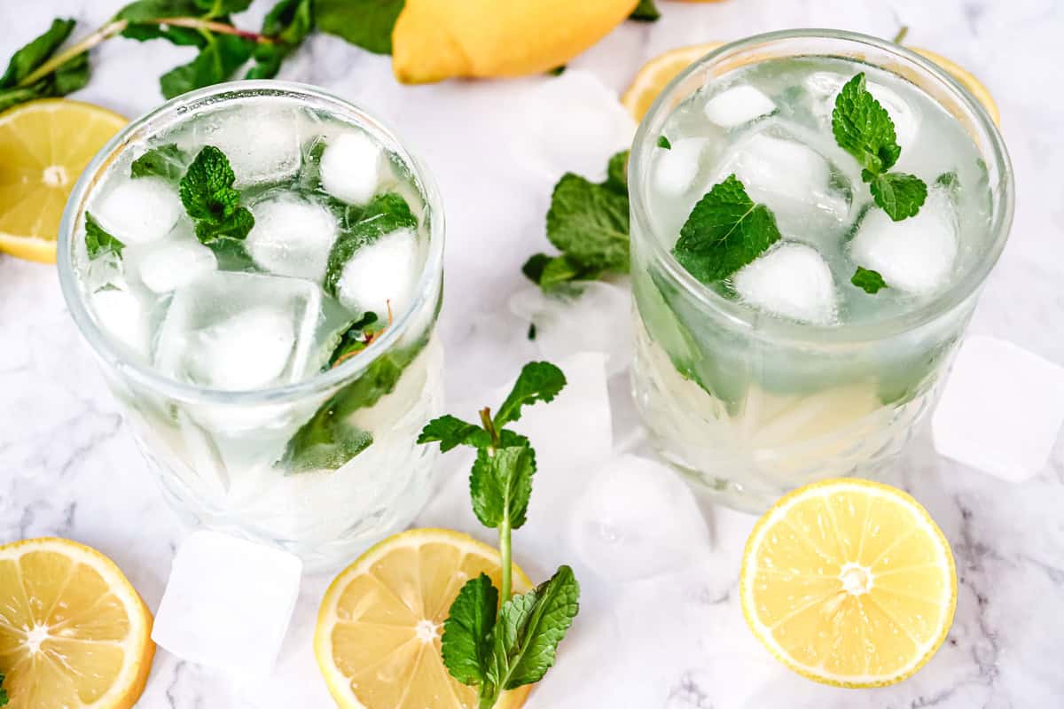 Two glasses of iced lemonade with mint leaves, surrounded by lemon slices, mint, and ice cubes on a white surface&mdash;an inviting Lemon Mojito Recipe perfect for any mocktail recipe collection.