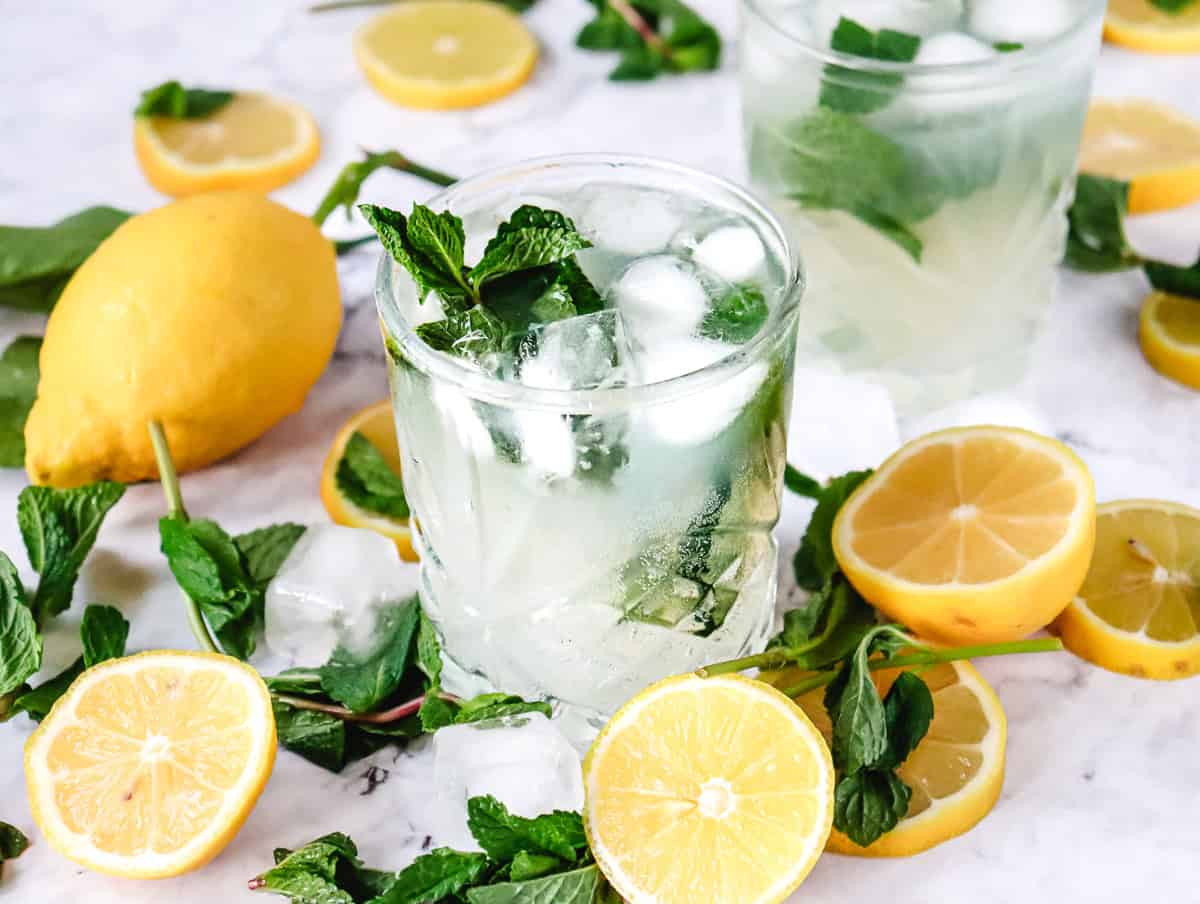 A refreshing Lemon Mojito Mocktail: a glass of iced lemonade with mint leaves, surrounded by lemon halves, mint sprigs, and ice cubes&mdash;perfect for your next mocktail recipe.