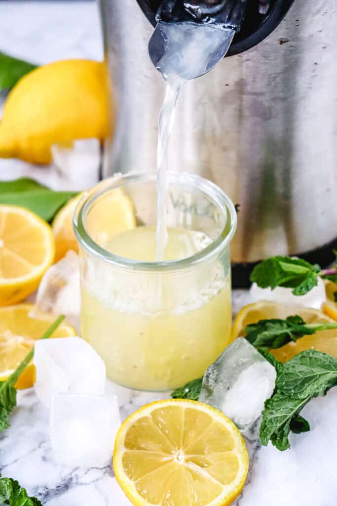 Fresh juice pours from a juicer into a glass, surrounded by lemons, mint leaves, and ice cubes&mdash;perfect ingredients for a refreshing Lemon Mojito Mocktail.