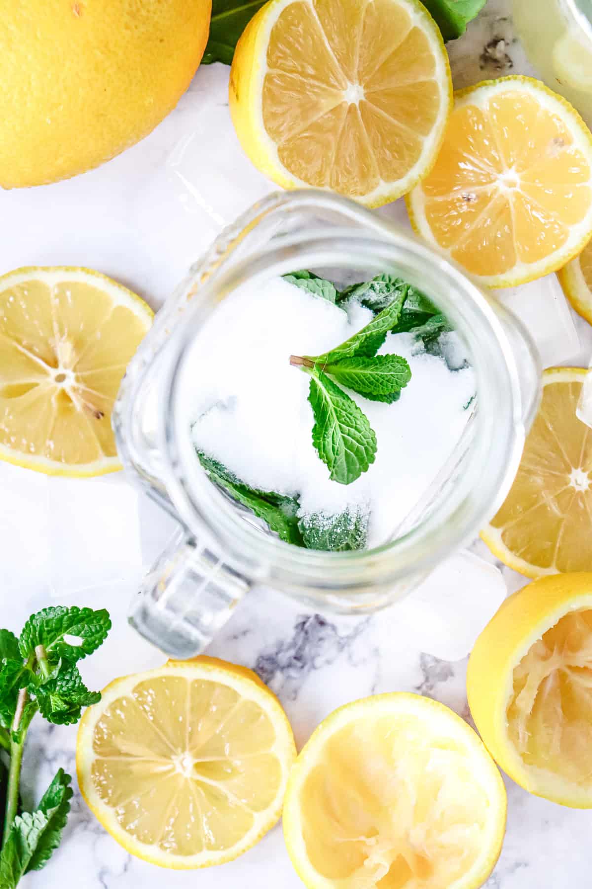 Top view of sliced lemons, mint leaves, and sugar in a glass jar on a marble surface&mdash;perfect ingredients for a refreshing Lemon Mojito Mocktail.