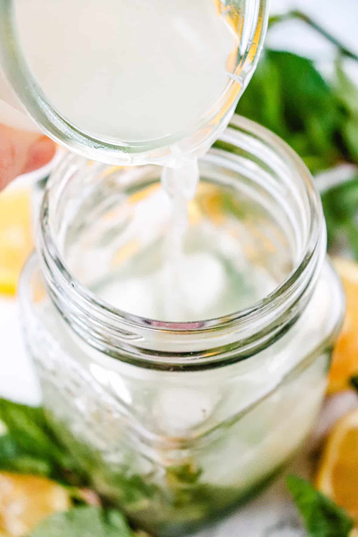 Lemonade being poured from a small pitcher into a glass jar, surrounded by lemons and mint leaves&mdash;perfect inspiration for a refreshing Lemon Mojito Mocktail.