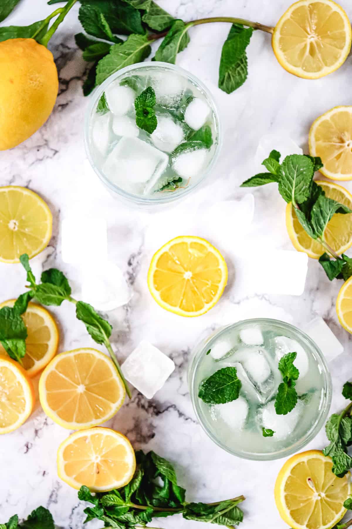 Two iced Lemon Mojito Mocktails with mint, surrounded by lemon slices, whole lemons, and mint leaves on a marble surface.