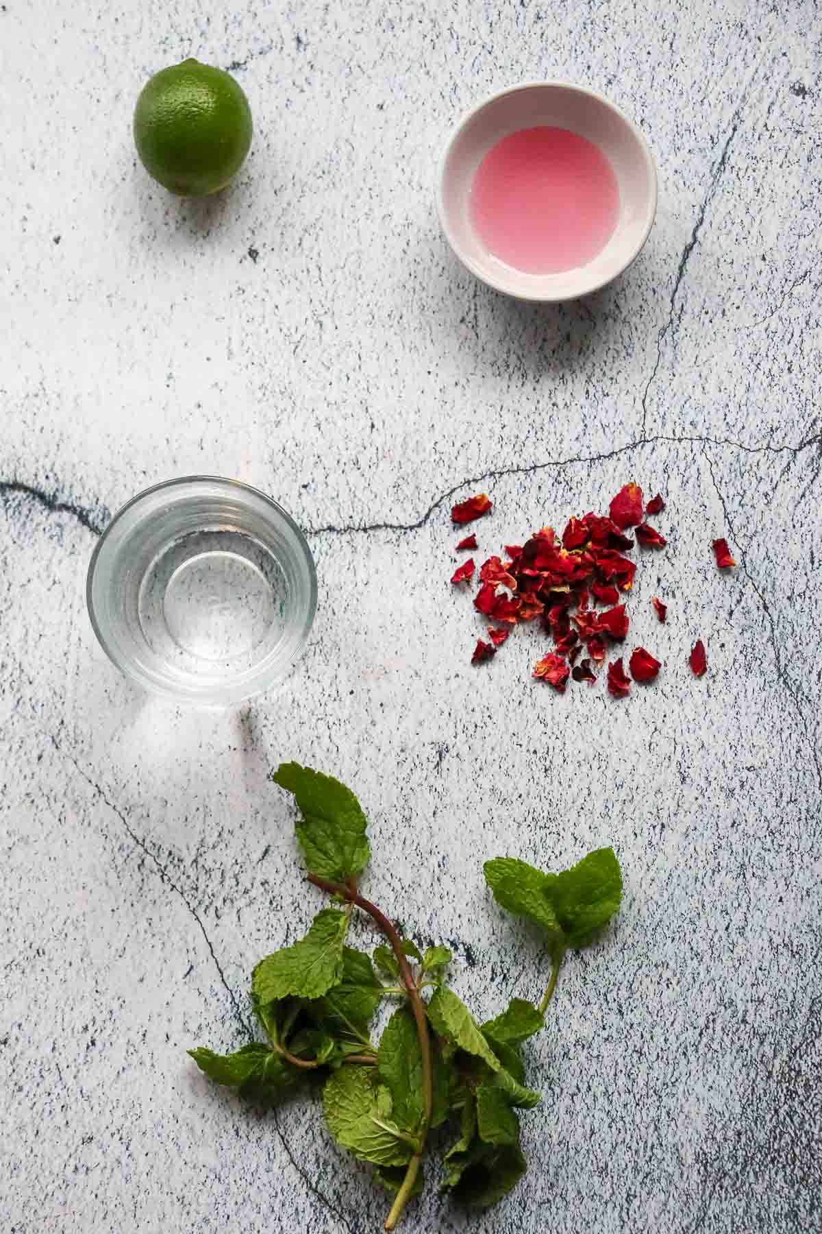 Lime, bowl of pink liquid, dried rose petals, mint leaves, and a glass of water on a light surface&mdash;perfect for crafting a refreshing non-alcoholic rose mojito mocktail recipe.