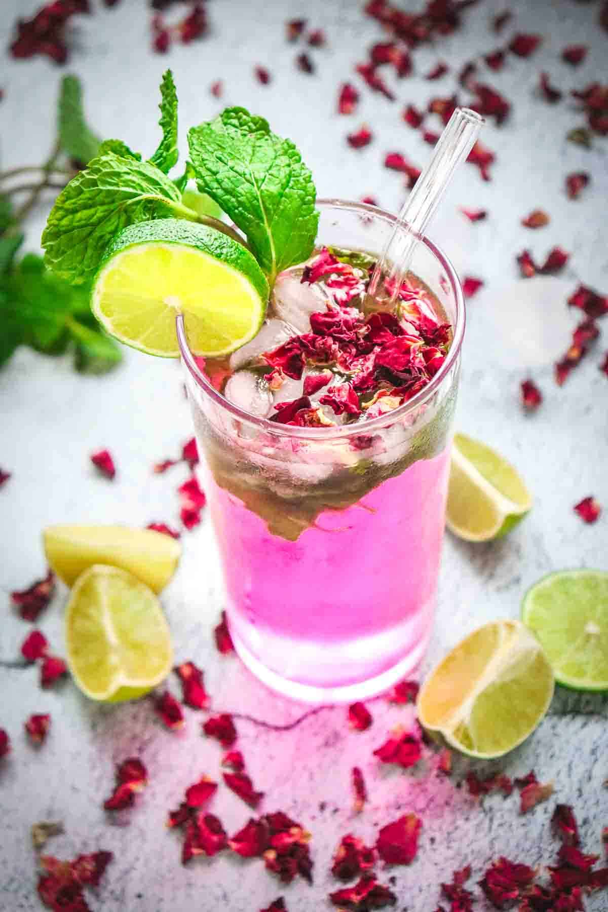 A pink non-alcoholic rose mojito mocktail recipe featuring ice, mint, lime slices, and rose petals in a glass, surrounded by lime wedges and petals.