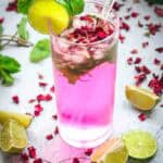 A pink non-alcoholic rose mojito mocktail served in a glass with ice, lime slices, mint leaves, rose petals, and a clear straw.