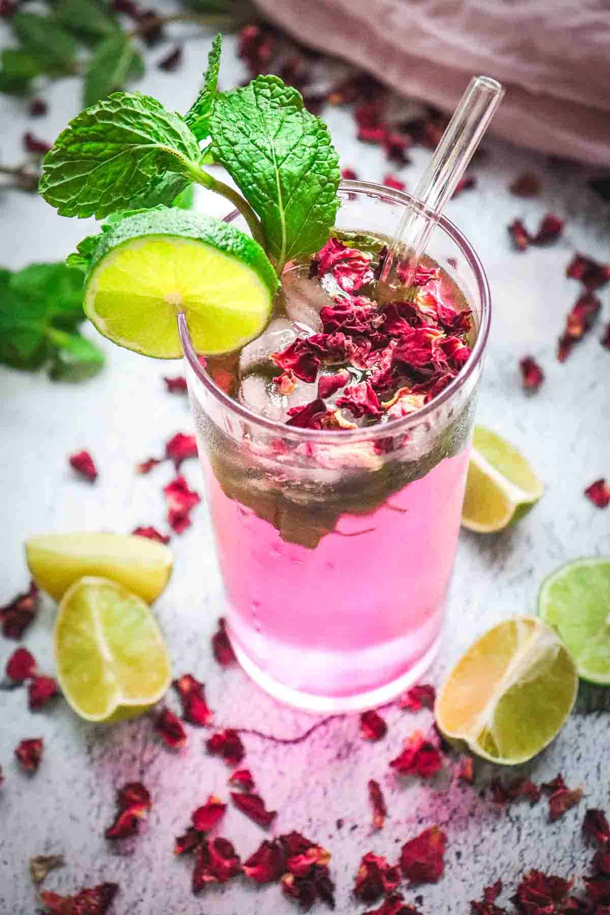 Tall pink non-alcoholic rose mojito mocktail with ice, lime slices, mint, and rose petals, surrounded by lime wedges and rose petals.