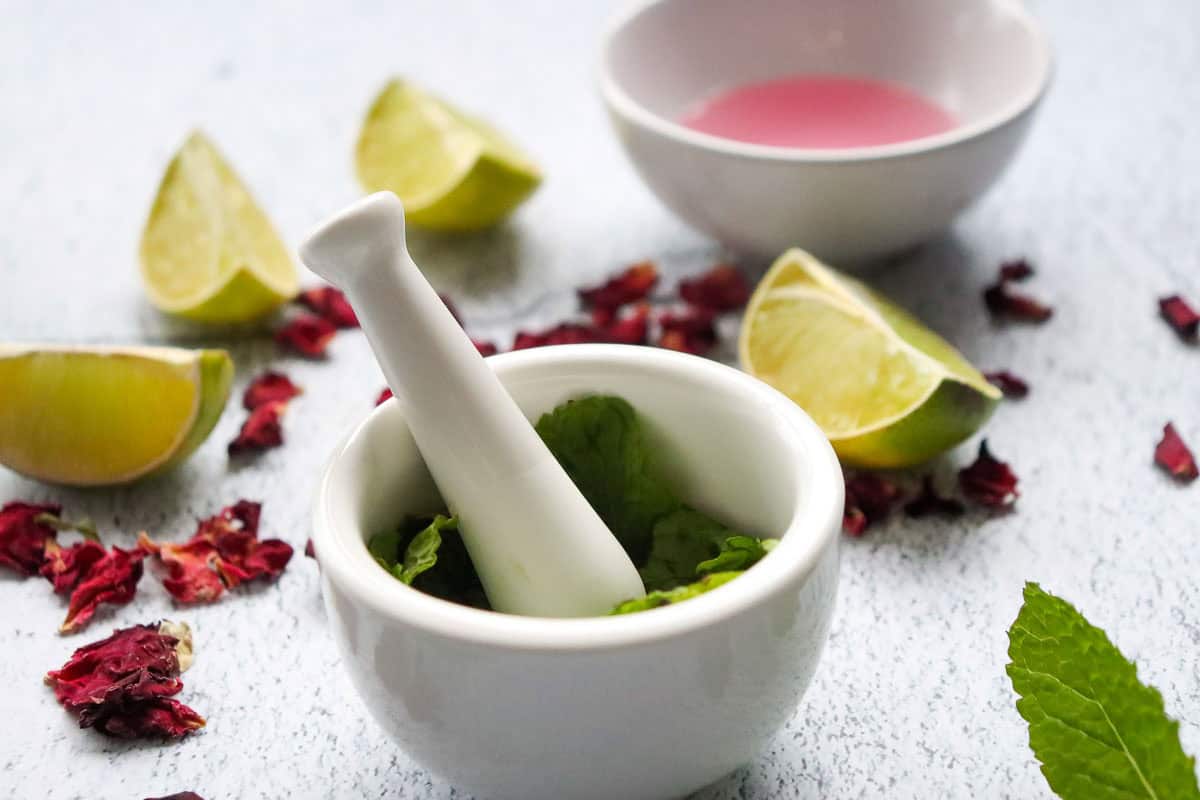 A mortar and pestle with mint leaves, lime wedges, dried rose petals, and a bowl of pink liquid&mdash;perfect for crafting a refreshing non-alcoholic rose mojito mocktail recipe.