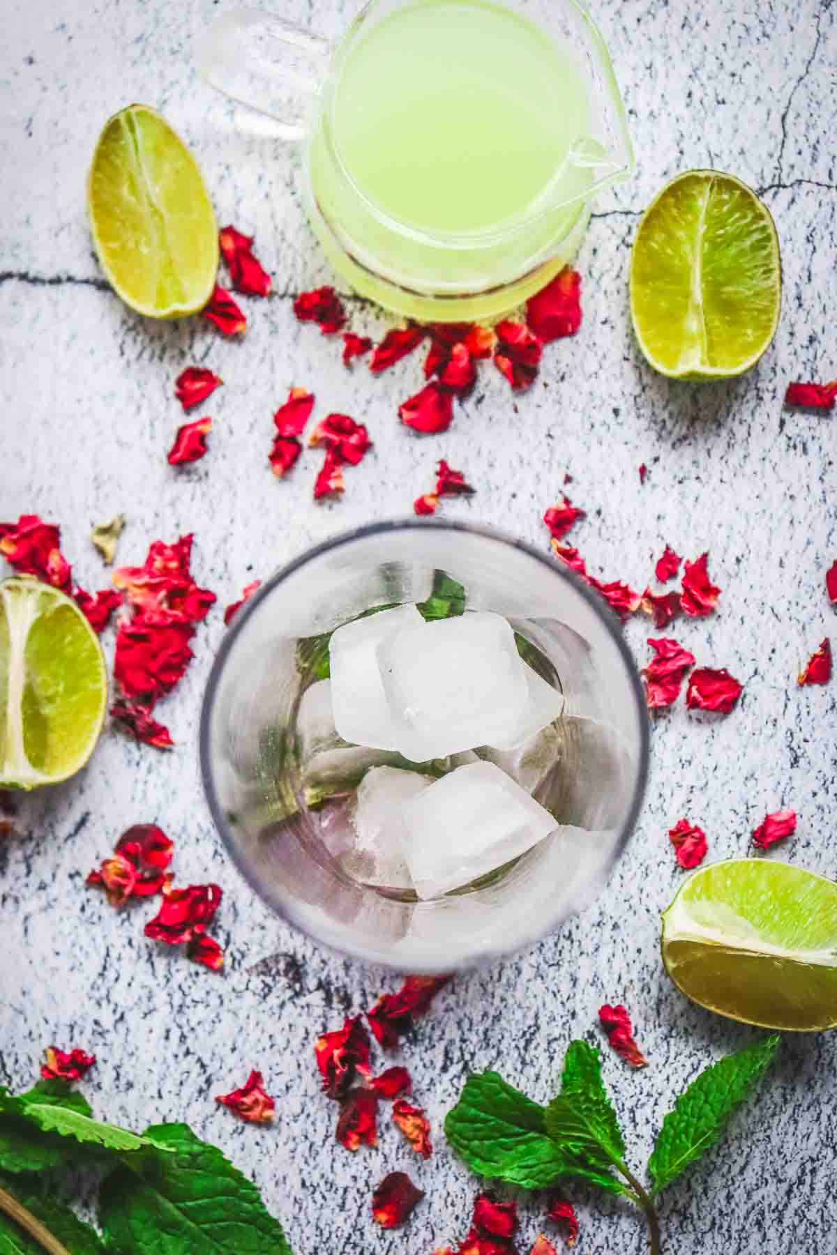 Top-down view of ice cubes in a glass, surrounded by lime halves, mint leaves, and red flower petals&mdash;perfect inspiration for a non-alcoholic rose mojito mocktail recipe.